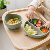 Sage green divided plate and bowl with healthy food on a highchair tray, baby's hand reaching in