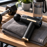 Black mini massage gun resting on a grey gym towel beside a dumbbell and water bottle